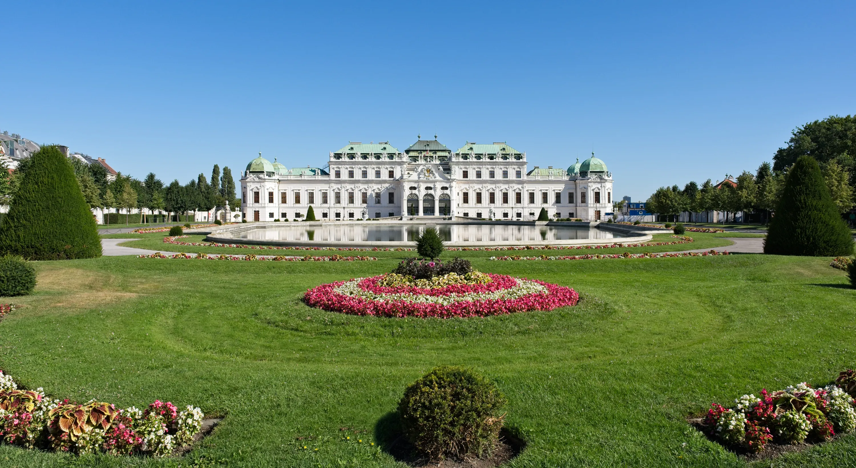 The Upper Belvedere in Vienna behind a reflecting pool, colourful flower beds and lawns beneath a clear blue sky.