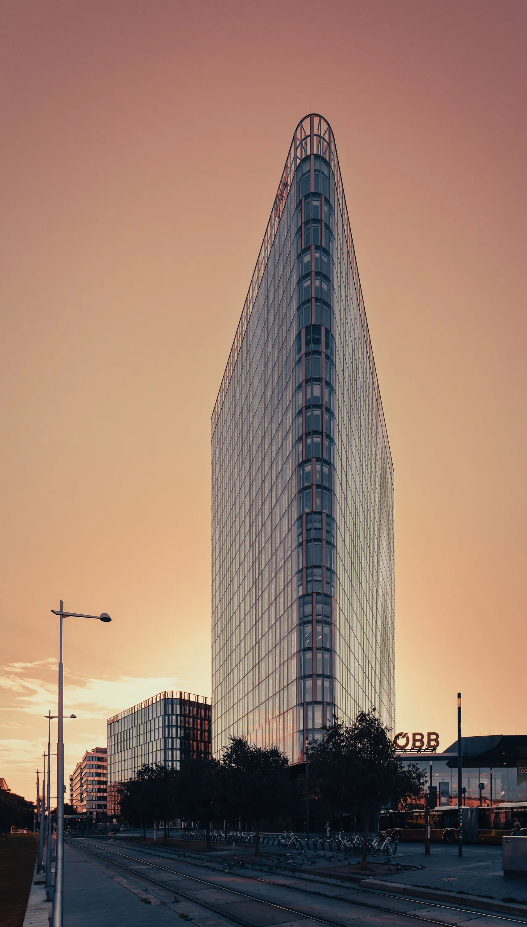 A tall glass building with a narrow, rounded corner rises against an orange-pink sky at Vienna Central Station; tram tracks, streetlights, bicycles and an ÖBB sign occupy the foreground.