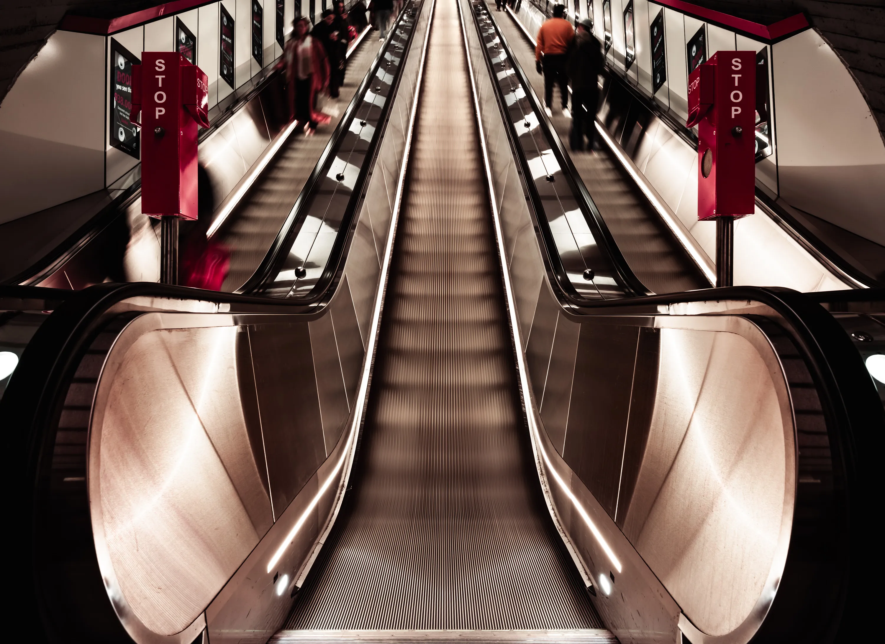 Three parallel escalators extend upward between illuminated metal balustrades at Karlsplatz underground station in Vienna. People appear blurred on the outer escalators, beside red emergency-stop columns.