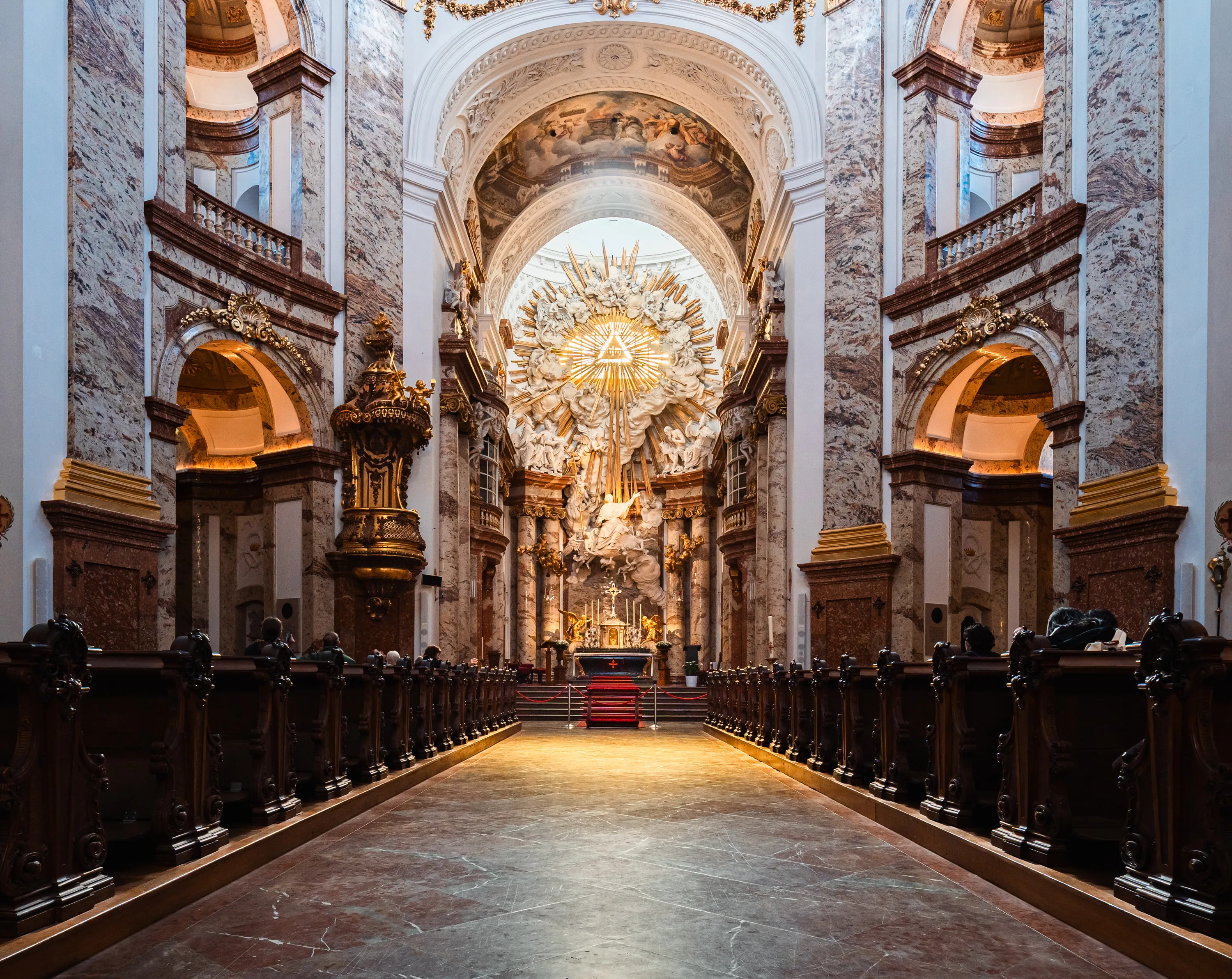 A central aisle between dark wooden pews in Vienna’s Karlskirche leads to an illuminated altar beneath a golden sunburst and sculpted figures, framed by marble-patterned pillars and painted vaults.
