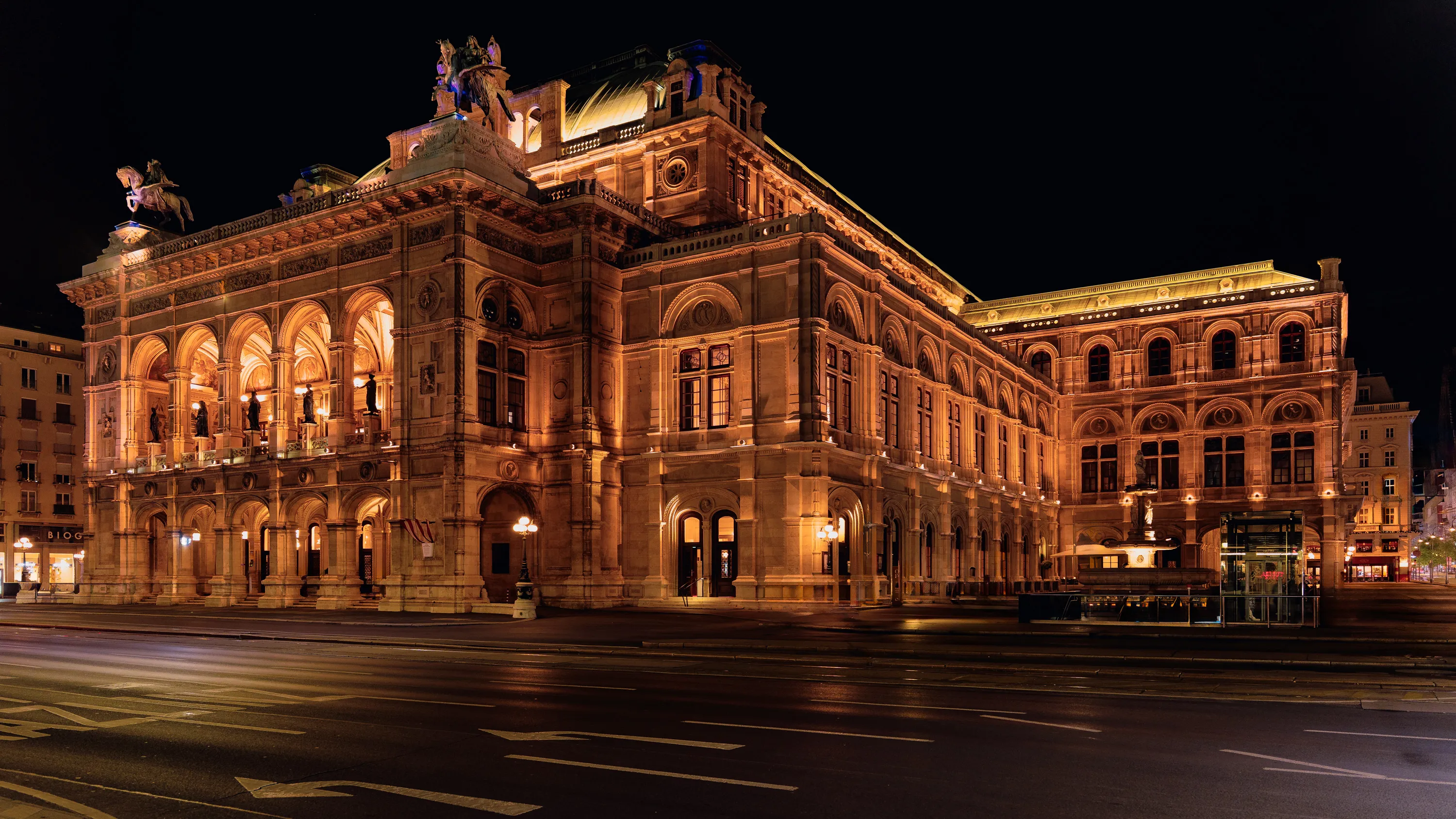 The Vienna State Opera glows in warm light against a black sky, with its arcades, roof sculptures and long side façade visible above faint traffic light trails.