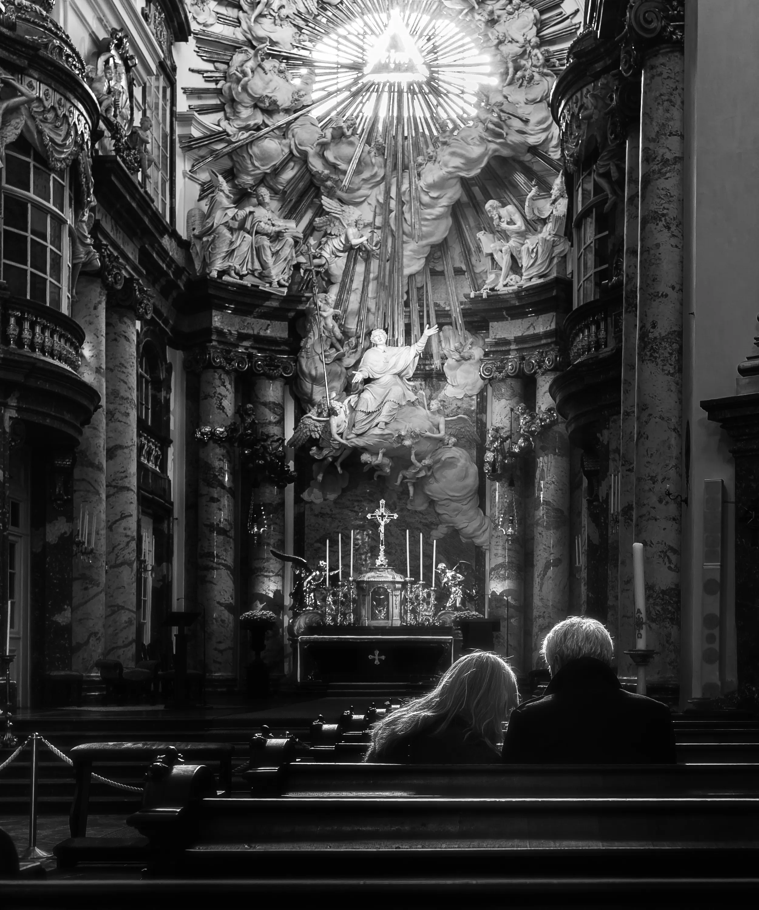In a black-and-white view inside Vienna’s Karlskirche, two people sit with their backs to the camera in a wooden pew, facing an altar with sculpted figures and a bright radiating ornament.